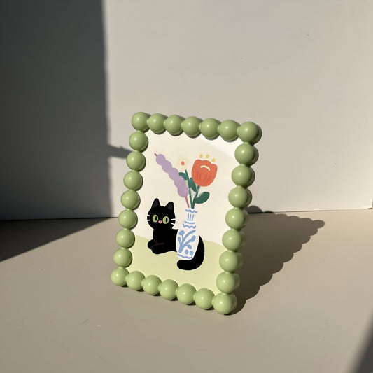 Green bead frame with a black cat and vase illustration on a neutral background